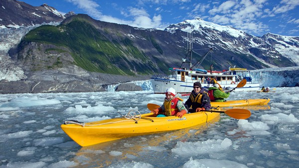 Travelers in a yellow kayak paddle in icy water with their Alaska small ship cruise seen behind them.