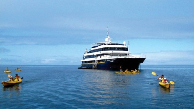 A mid-sized Galapagos ship shown with three kayakers in front in yellow kayaks.