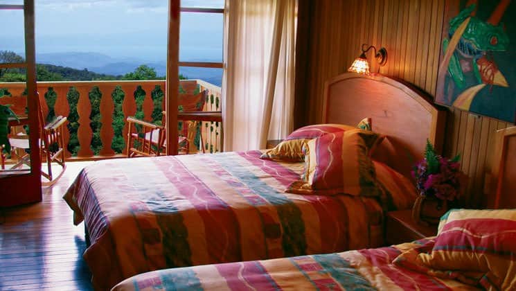 Room interior with two beds and balcony with two chairs and rainforest views at Hotel Fonda Vela in Monteverde, Costa Rica