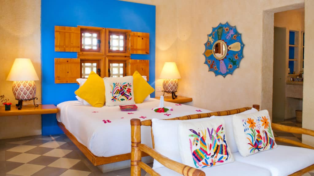 Large bed covered in red flower petals and colorful pillows, with loveseat and Mexican-style artisan wall hangings at Casa Natalia in San Jose del Cabo on the Baja Peninsula