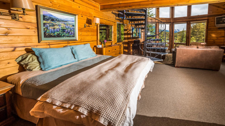 Inside the Eagle's Nest Cabin with a wood interior, cozy decor, king-sized bed, spiral staircase leading to the loft and large windows, at the Tutka Bay Wilderness Lodge in Alaska