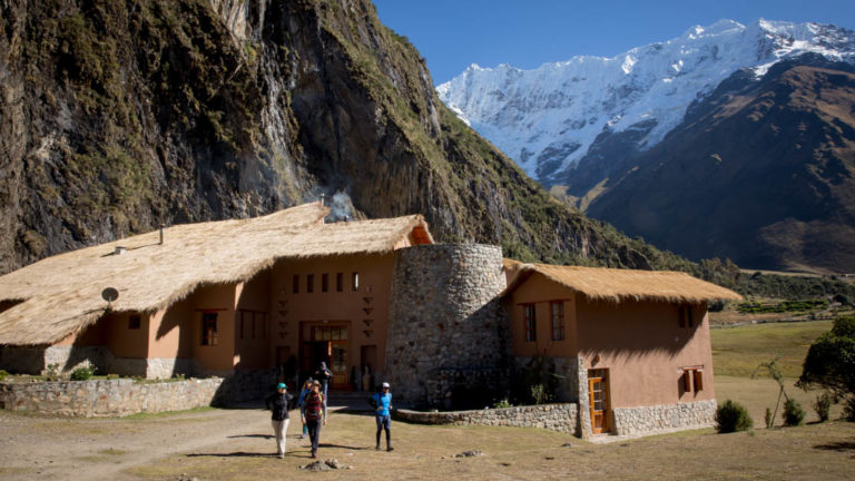 A group of trekkers stand outside the Salkantay Lodge, Adventure Resort, one of four hotels that are part of the Mountain Lodges of Peru on the Inca Trail to Machu Picchu. The building has thatch roofs and stands beneath a towering mountain in the Andes.