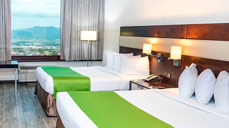 A window with a view of San Jose, Costa Rica, and two beds with white linens and a green duvet inside the standard double room at the park inn by radisson.