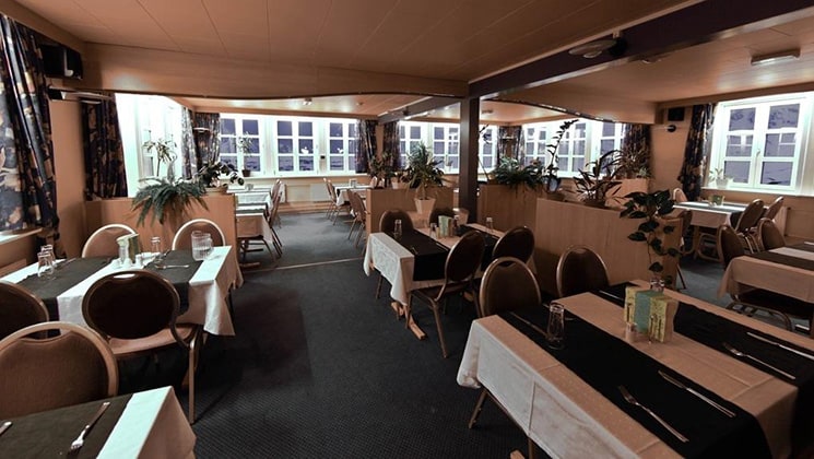 Dining room of Hotel Kulusuk in Greenland, with tables covered in white tablecloths & black runners, flanked by basic banquet chairs.