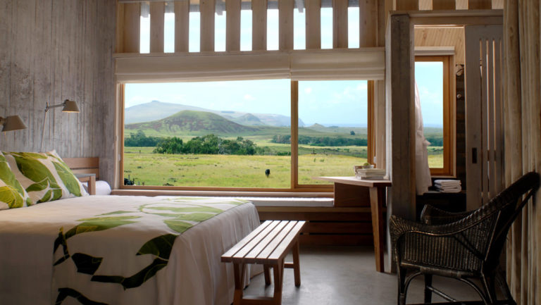 A room with a large window overlooking the mountains and green fields on Easter Island, with a full bed and modern furniture, at Posada de Mike Rapu, a luxury eco hotel.