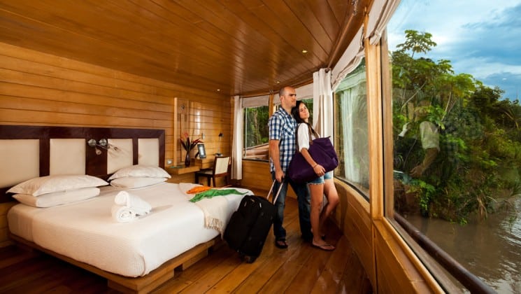 Man and woman holding luggage gaze out picture window in Master Suite with large bed aboard Delfin II riverboat on Amazon River cruise