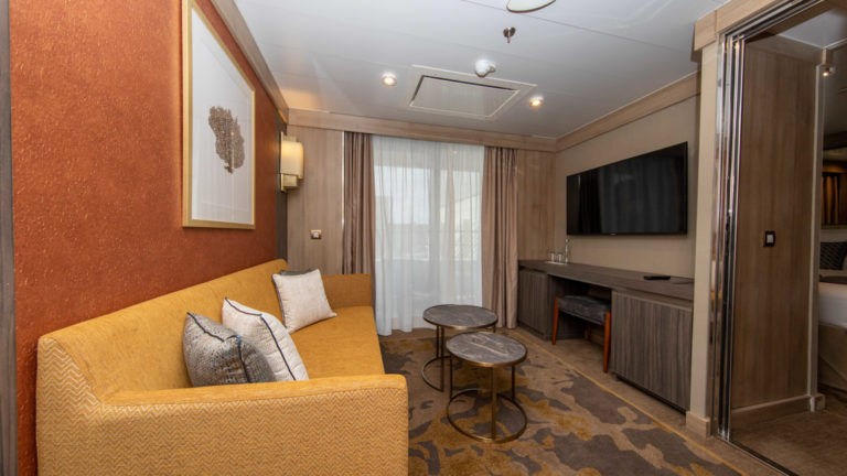 Seating area of Superior Suite with golden-colored couch, coffee tables, wet bar, TV and balcony aboard polar small ship World Explorer.