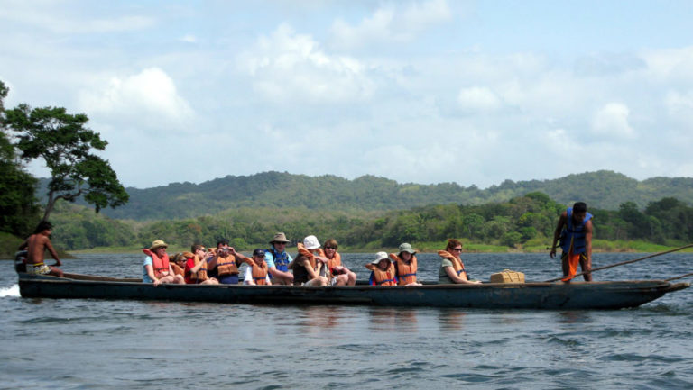 a group of adventure travelers rows a dugout canoe on flat water with mountains in the background on a sunny day during the best of panama land tour
