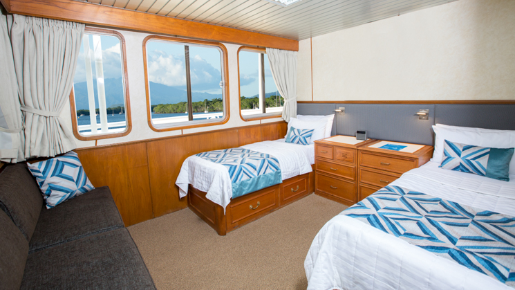 Deluxe Stateroom with two beds, bedside tables, a couch and three large windows aboard Coral Expeditions II in Australia