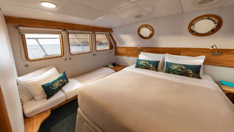 Double bed with tan bedding & decorative pillows, flanked by bedside bench set up for sleeping, aboard Corals Galapagos yachts.
