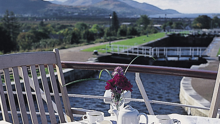 Lord of the Glens deck with a table and chair set up for tea with the locks behind the small ship cruise in Scotland.