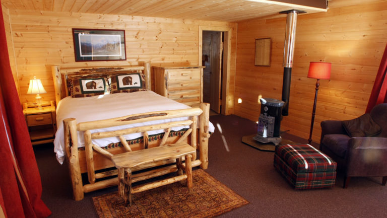 The interior of a rustic cabin with a full bed, wood-crafted furniture, reading lamps, a wood stove, and a private bathroom at Winterlake Lodge, an Alaska resort recognized by National Geographic for its wilderness experience