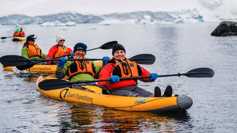 Polar travelers pose for the camera while paddling an inflatable yellow kayak among snowfields in calm water.