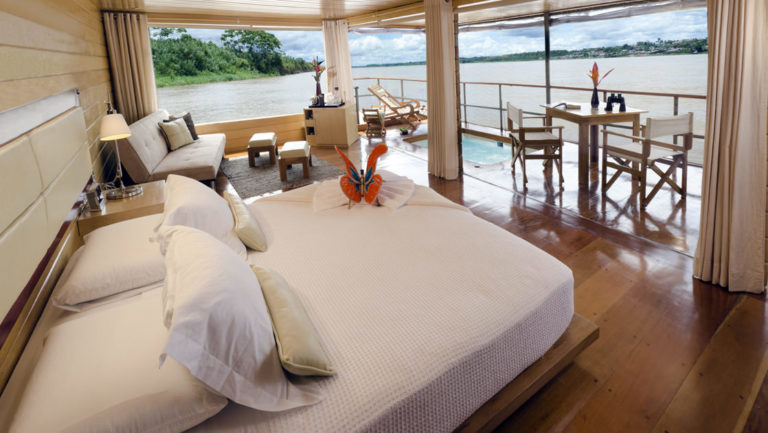 Inside a Deluxe Suite with bed, couch and private balcony with table, chairs, Jacuzzi and lounge chair aboard Delfin I on the Amazon River