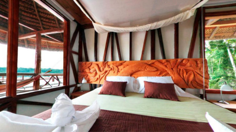 A king-sized bed with fresh maroon and white linens is made in a room displaying traditional Ecuadorian architecture and woodwork at the Napo Wildlife Center, a luxury eco lodge surrounded by a rainforest biosphere reserve the Amazon.