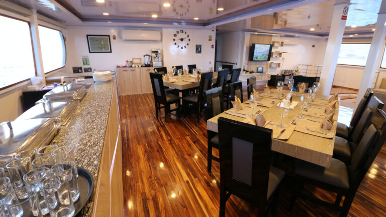 Dining area of Petrel boat in Galapagos with wood flooring, granite bar, large view windows, white walls & two 8-top tables.