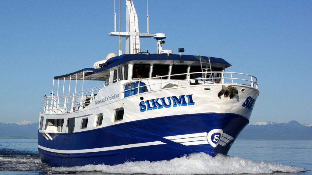 Sikumi small Alaska ship cruises in calm water on a sunny day, showing her dark blue & white exterior, main deck, wheelhouse & antennae.