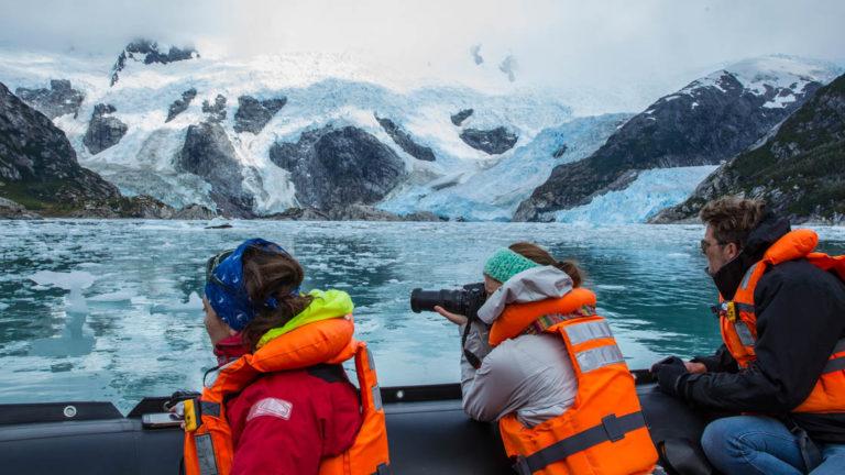 adventure travelers sitting on the edge of a small ship taking pictures across the water of patagonia fjords