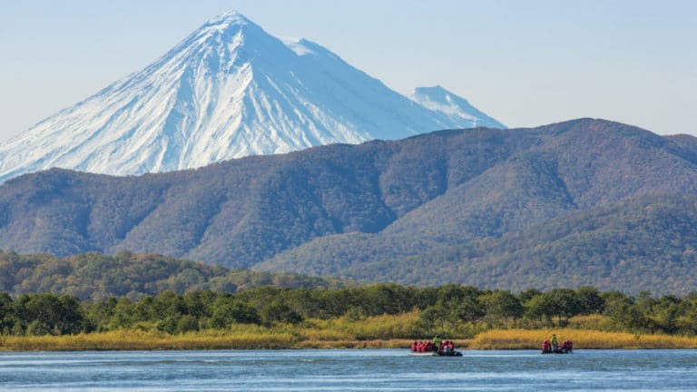 three groups of travelers take kodiac rides on the Zhupanova River, Russia. A jagged snow mountainrnage in the distance behind a lower forested mountain, seen on Across the Bering Sea Cruise