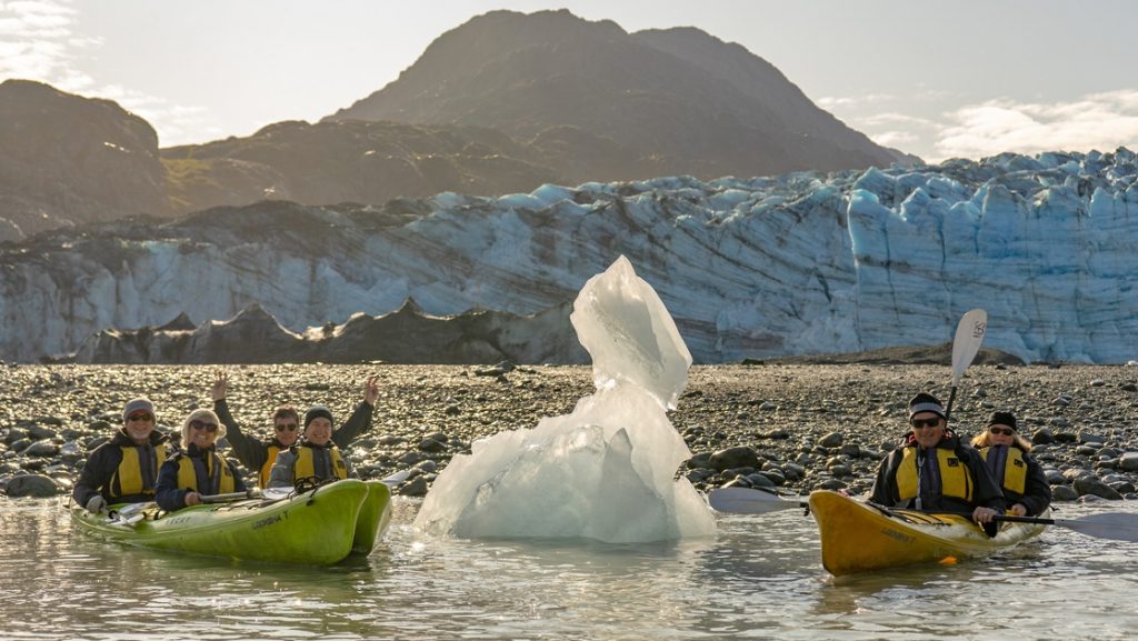 In Alaska a group of kayakers float in green and yellow kayaks next to a white iceberg and in front of an icy teal glacier.