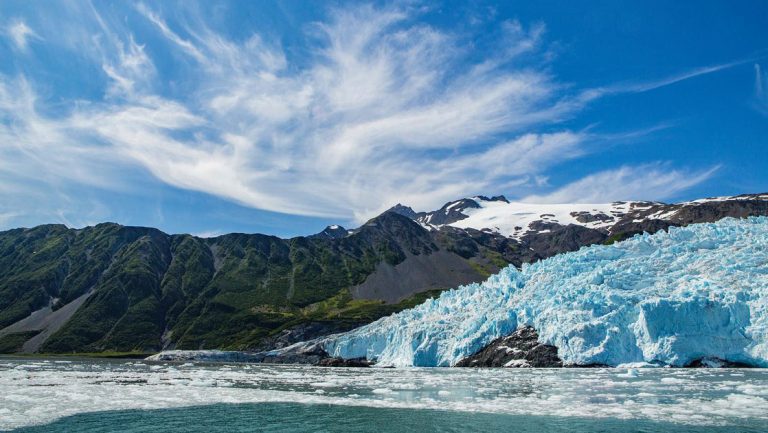 Icy blue glacier leads into a body of water beside green mountains under a blue sky on the Alaska Grand Adventure.