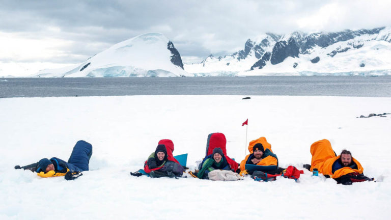 travelers lay on the ground in sleeping bags in antarctica with the ocean behind them and mountains in the distance