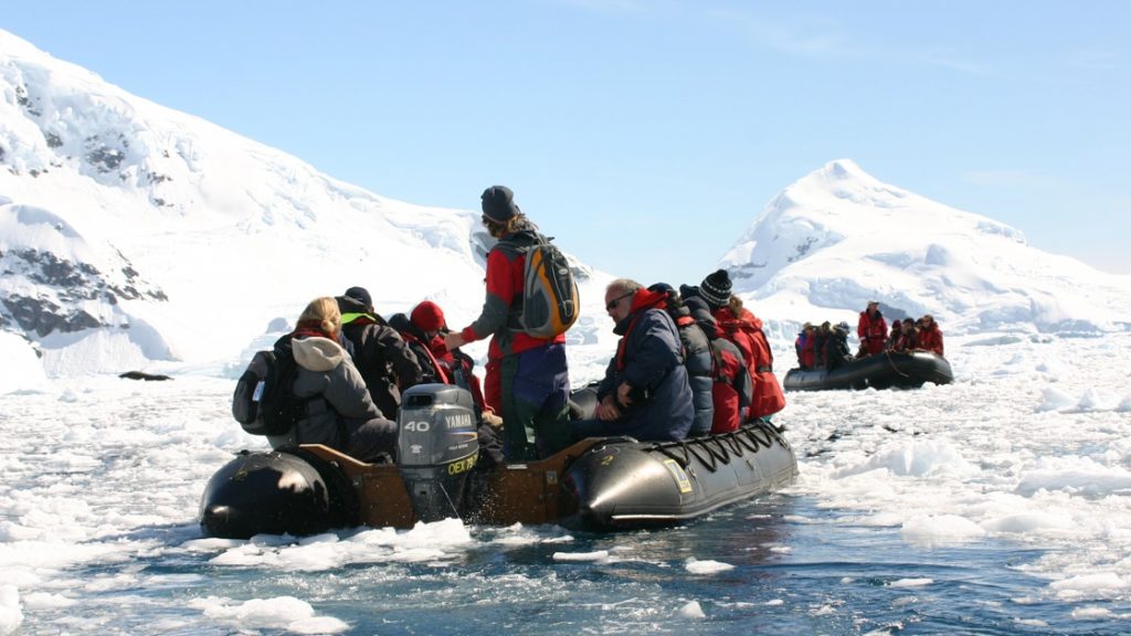 Antarctica travelers in jackets of various bright colors sit in a Zodiac as it cruises amongst icebergs on a sunny day.