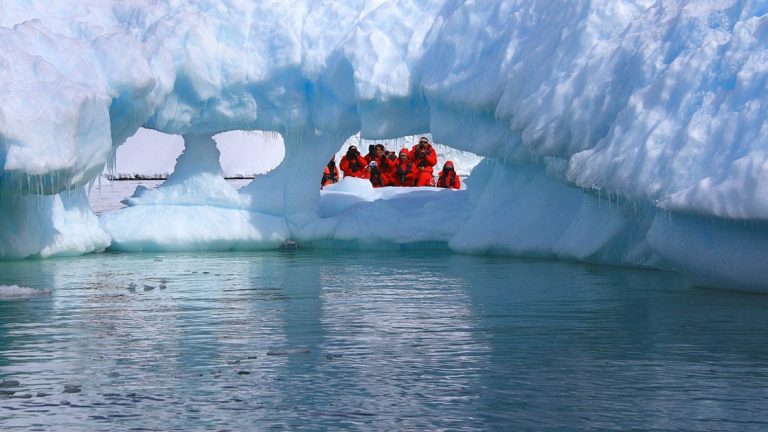 Group of travelers on a small expedition ship in Antarctica wearing their red parkas on a zodiac excursion and looking through one of several holes in an iceberg.