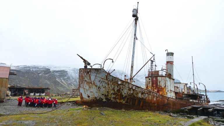 Group of travelers wearing red parks on a land excursion looking up at an abandoned rusty ship in Antarctica.