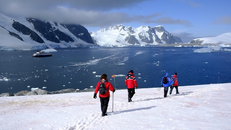 Antarctic travelers walk along a prescribed path in the snow with calm waters and their expedition ship in the background, on a sunny day during the Whale Science Voyage.