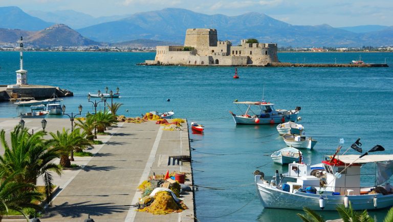 Turquoise sea with a beige castle in the background & paved promenade along the water in the foreground, in Nafplion, Greece.