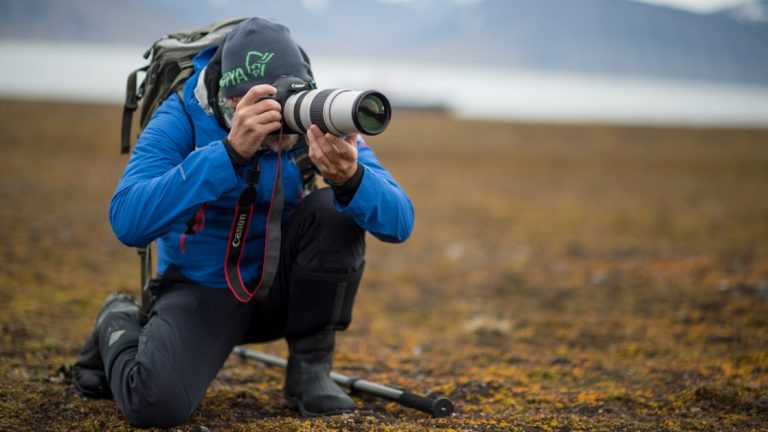 Male photographer in bright blue jacket kneels down by green and gold tundra to take a photo.