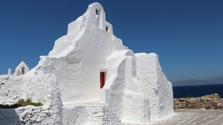 Stucco, bright white chapel with red door sits beside deep blue ocean, seen on the Best of Greece & Turkey small ship cruise.