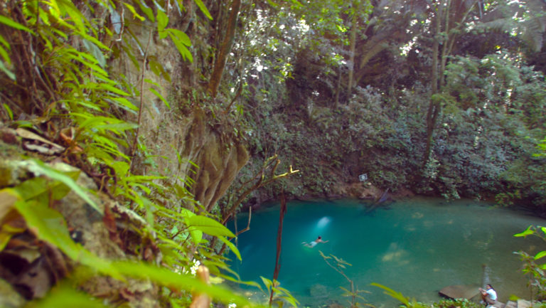 The Blue Hole in Belize with a swimmer in the turquoise water and green foliage surrounding it.