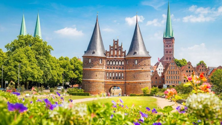Brick castle with round pointy tops beside bright green grass with purple flowers in the foreground on a sunny day in Germany.