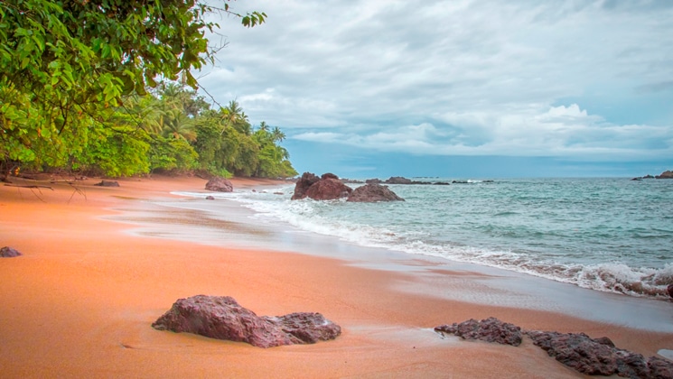 Pink-sand beach with red rocks, green forest & turquoise waves under a cloudy sky in Costa Rica.