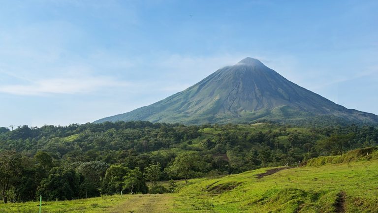 Arenal volcano from afar with bright green forest & grassland in the foreground, on a sunny day in Costa Rica.