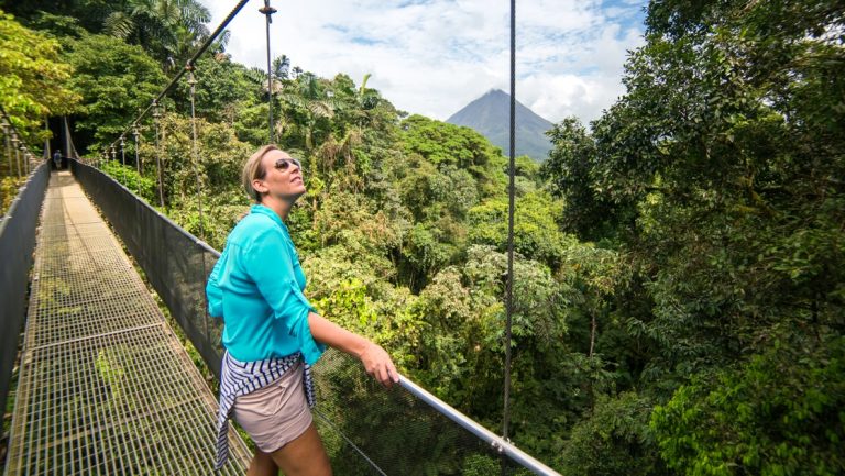 Woman in beige shorts & teal shirt stands on a hanging bridge in the Costa Rica rainforest & looks up into the trees on a sunny day.