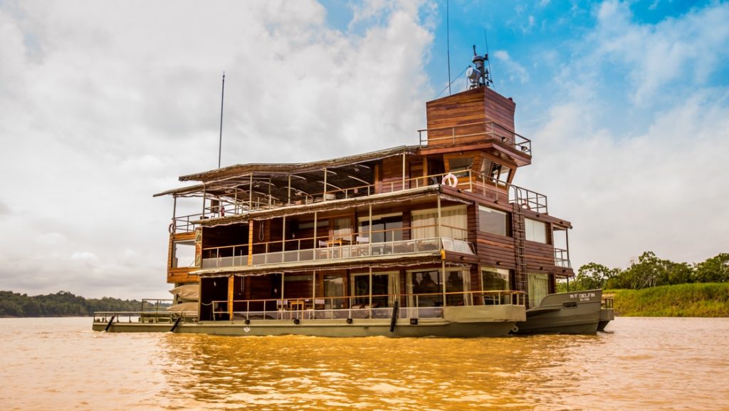 Delfin I Amazon river boat cruises through muddy water on a sunny day, with 3 wooden decks & 2 sage green metal hulls.