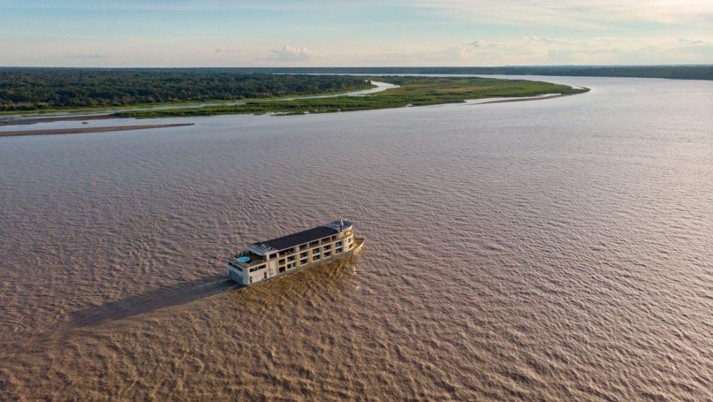 Aerial view of modern beige riverboat with 3 decks cruising in the middle of the large Amazon River on a clear day in Peru.