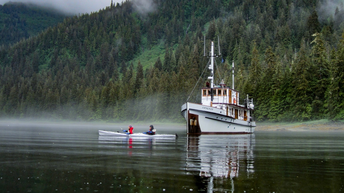 Against a green forested hillside, a small white wooden ship, Catalyst, floats in dark green water as guests in a double kayak paddle around it.