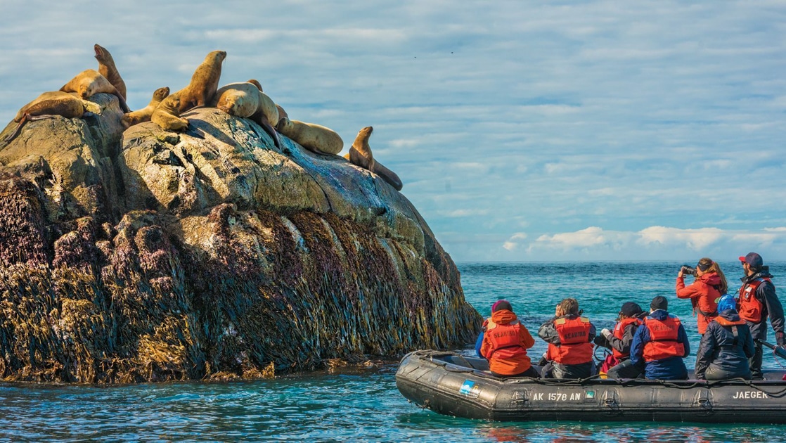 Alaska travelers sit & stand in black Zodiac boat while photographing seals atop a tall rock rising out of the water in a cloudy day.