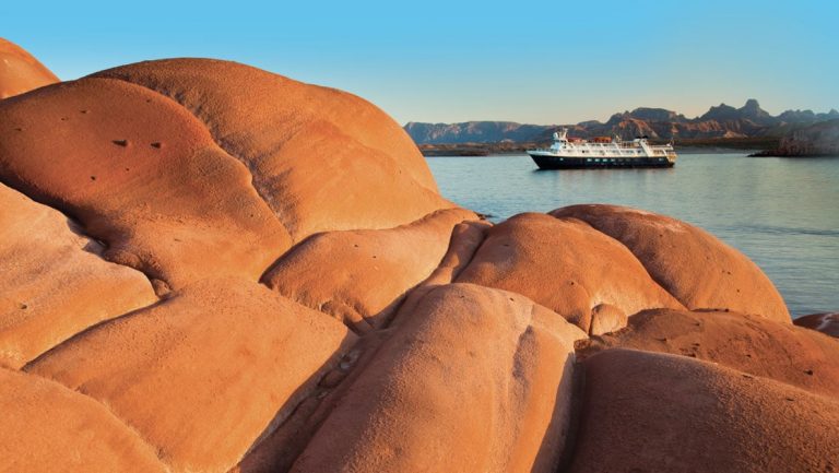 Sandstone rock in foreground with a small expedition ship in the background on a sunny day in Baja during a Sea of Cortez small ship cruise.