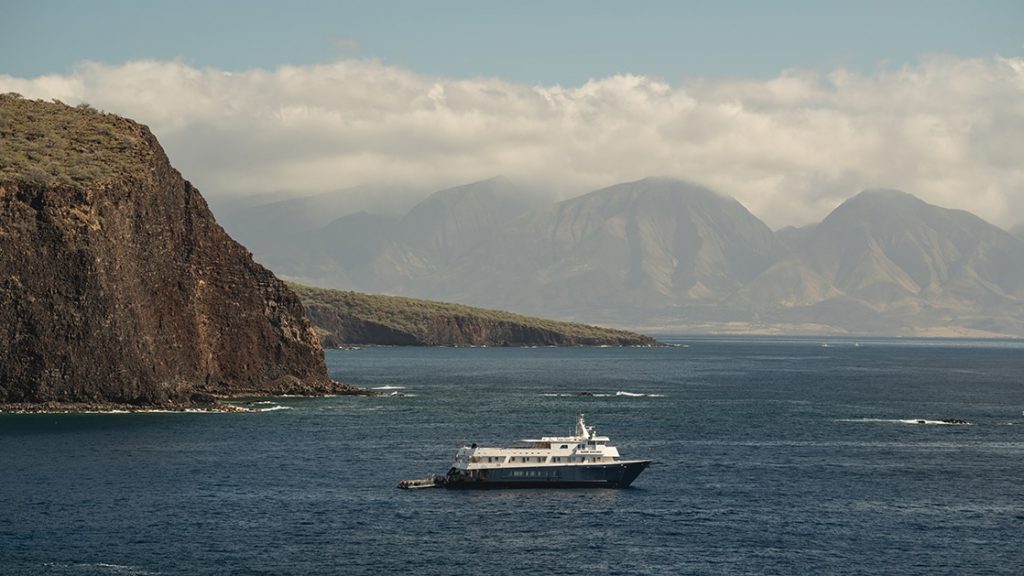 A small Hawaii yacht seen cruising off the coast of Maui sailing away from the mountainous coastline.