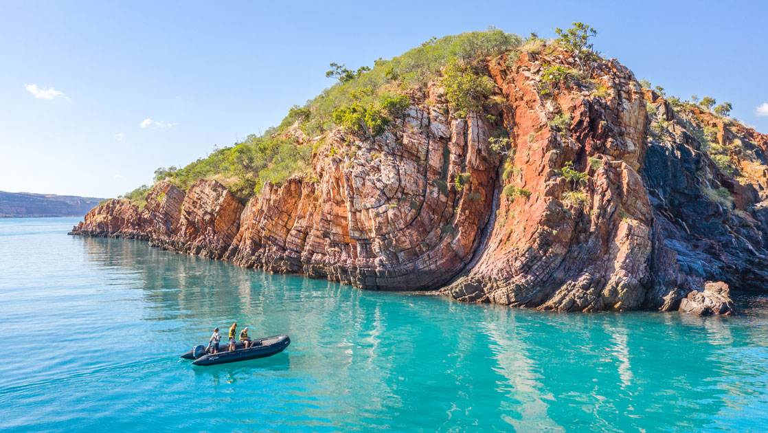 Zodiac of people pulls up to rocky red islet with teal-colored water & bright green plants in Australia's Kimberley region.