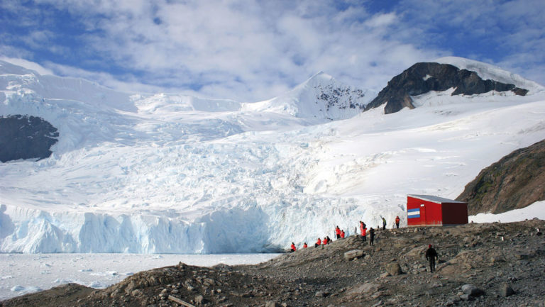 travelers hiking on an antarctica shore with a red hut next to them and a giant white iceberg behind them