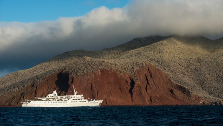 The all white Galapagos small ship the Legend floats in the dark blue ocean against a deep red hillside of Rabida island.