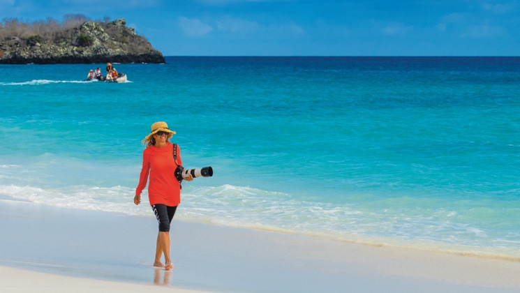 Woman in red shirt & black shorts walks a white-sand beach beside turquoise water on the Wild Galapagos Escape Cruise.