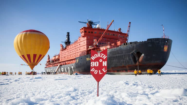 parked on the ice behind a red arrow marking the true north pole the large 50 years of victory ship sits, next to it a yellow and orage hot hair balloon is inflated
