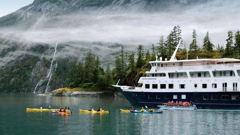 In front of tall fjord walls with waterfalls the white and blue Safari Endeavour ship floats as a group of kayakers paddle yellow and blue kayaks.